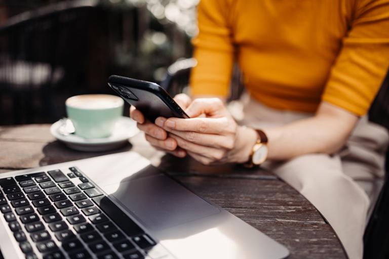 Person holding a smart phone while sitting at a table with a laptop